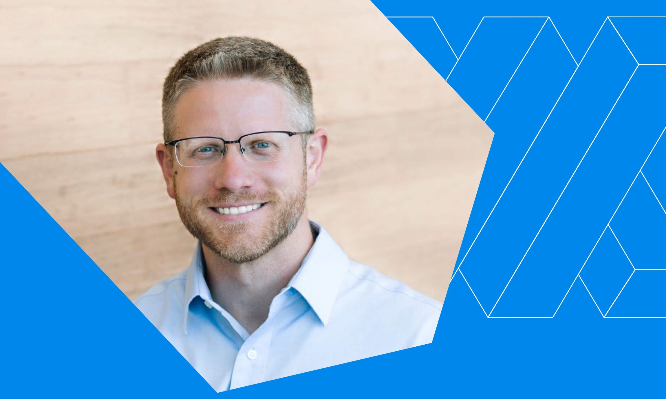 Portrait of a smiling man with short light brown hair, glasses, beard, and light blue shirt against a wooden background with blue geometric overlay.